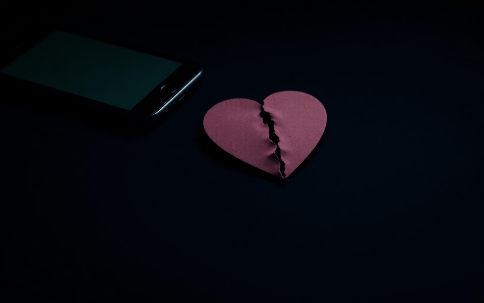 A torn paper heart on a dark surface beside a faintly lit smartphone, somber and minimal