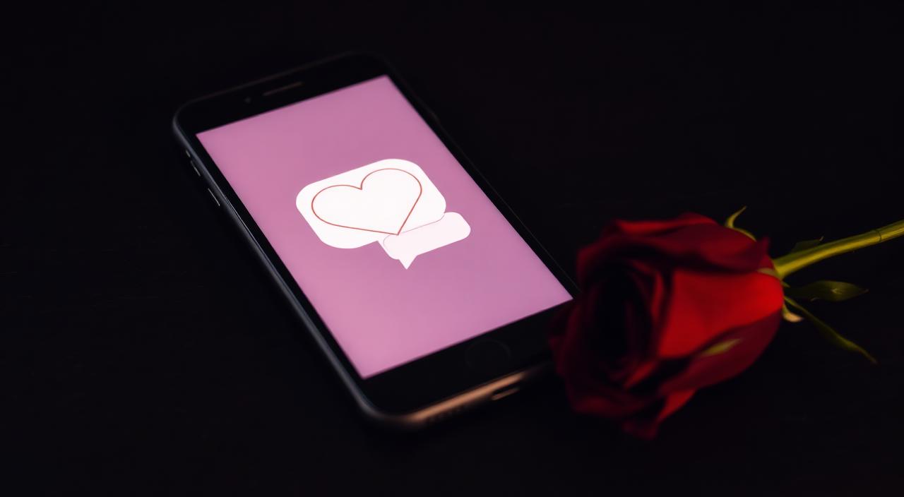 Smartphone glowing softly with a heart icon and message bubble beside a single red rose on a dark surface